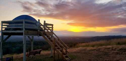 A wooden platform with a blue dome structure overlooks a sunset, casting warm colors across the sky and landscape.