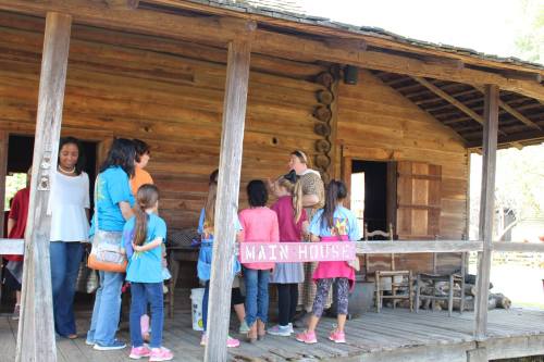 A group of children and adults gather on the porch of a rustic wooden building labeled "Main House."