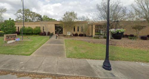 A brick building with a landscaped entrance, featuring a sign and trees, under a cloudy sky.
