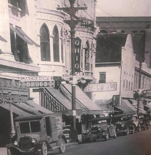 Historic street scene featuring vintage cars, storefronts, and a hotel sign, capturing early 20th-century architecture.