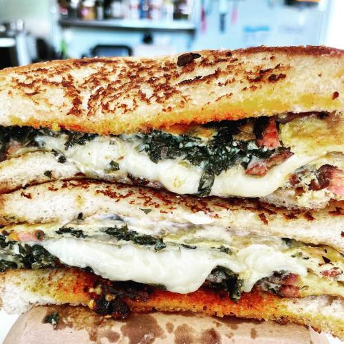 A close-up of a grilled sandwich with melted cheese, greens, and layers of meat, toasted to a golden brown.
