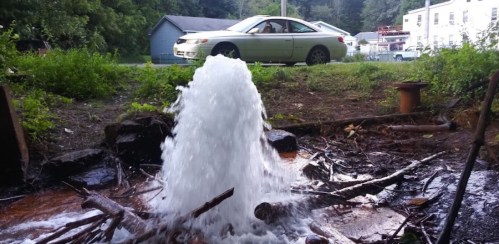 A water fountain erupts from the ground, with a car parked nearby and greenery in the background.