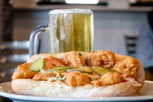 A close-up of a shrimp po' boy sandwich with pickles and a frosty mug of beer in the background.