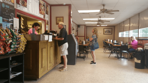 A casual dining area with tables, a counter, and customers ordering food. Snacks are displayed on a shelf.