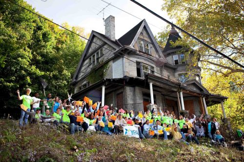 A group of people in bright shirts gather in front of an old, abandoned house, holding signs and smiling.