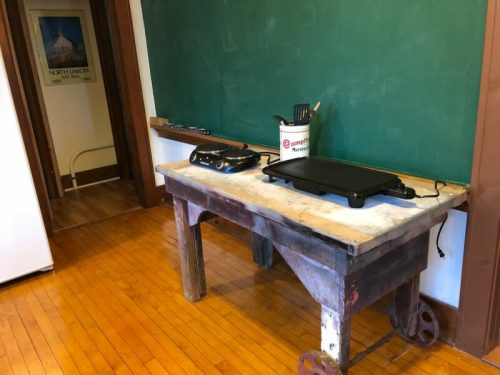 A wooden table with a griddle on top, next to a chalkboard in a room with wooden floors.