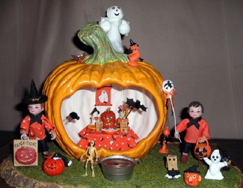 A whimsical Halloween scene featuring a pumpkin house with dolls, ghosts, bats, and festive decorations.