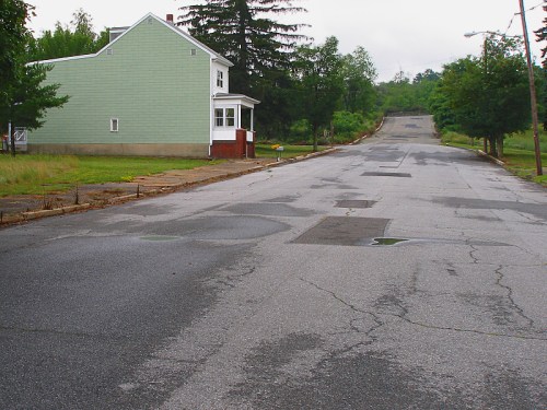 A quiet, overgrown street with a green house on the left and a sloped road leading into the distance.