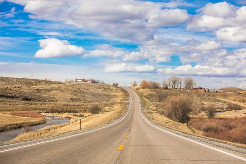 A winding road stretches through a rural landscape with rolling hills, sparse trees, and a blue sky dotted with clouds.