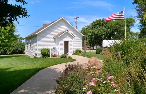 A white historic schoolhouse with a flag, surrounded by green grass and colorful flowers on a sunny day.