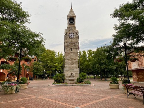 A tall clock tower stands in a park surrounded by trees and flower planters, with a cloudy sky above.