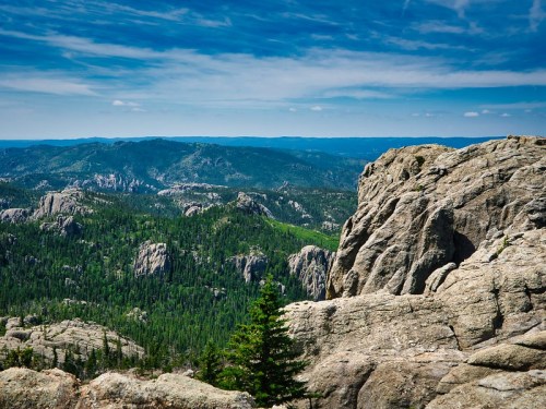 A panoramic view of rugged mountains and lush green forests under a blue sky with scattered clouds.