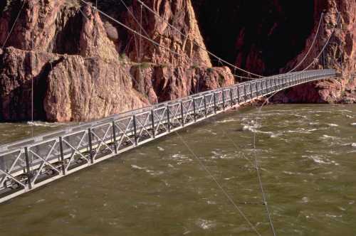 A suspension bridge spans a river, surrounded by steep rocky cliffs and turbulent water below.