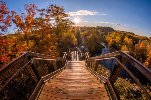 A wooden staircase leads down to a river, surrounded by vibrant autumn foliage and a sunset in the background.
