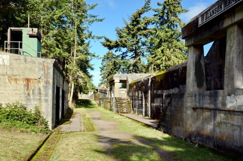 Abandoned military fortifications surrounded by trees under a clear blue sky, with pathways and stone structures visible.