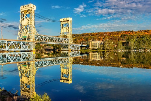A yellow lift bridge reflected in calm water, surrounded by autumn foliage and a clear blue sky.