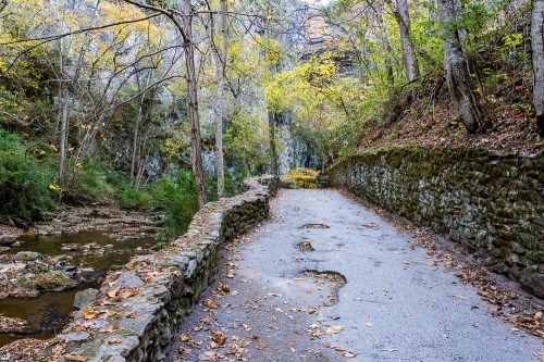 A serene pathway lined with stone walls, surrounded by trees with autumn leaves and a gentle stream nearby.