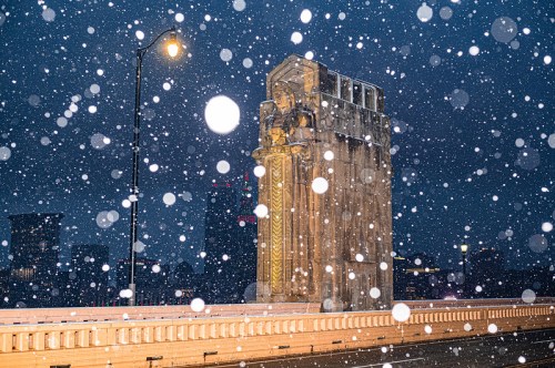 A snow-covered bridge at night, featuring ornate pillars and soft streetlight glow amidst falling snowflakes.
