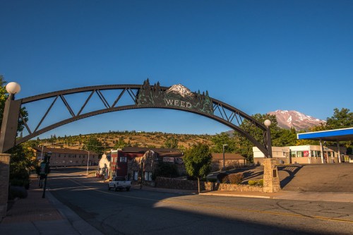 Archway sign reading "Weed" with mountains in the background and a clear blue sky. Quiet street scene.