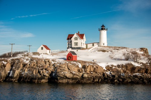 A snowy landscape featuring a white lighthouse and red-roofed buildings on a rocky shore under a clear blue sky.
