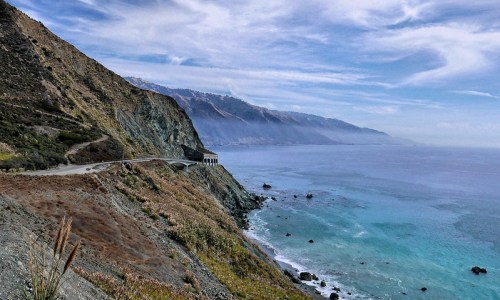 Scenic coastal view with a winding road along cliffs, overlooking the ocean and distant mountains under a cloudy sky.