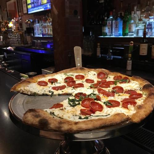 A pizza with fresh basil and cherry tomatoes on a metal serving tray, set against a bar backdrop.