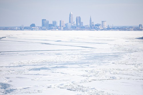 A snowy landscape with ice covering a lake, featuring a distant city skyline under a clear sky.