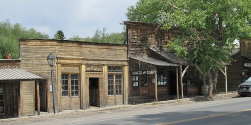 A row of rustic wooden buildings in a historic town, with trees and a road in the foreground.