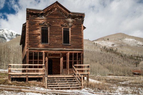 Abandoned wooden building with a steep roof, surrounded by mountains and trees under a cloudy sky. Snow on the ground.