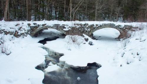 A stone bridge arches over a snowy stream, surrounded by trees in a winter landscape.