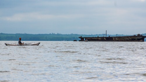 A person in a kayak paddles near a rusted shipwreck on a calm body of water under a cloudy sky.