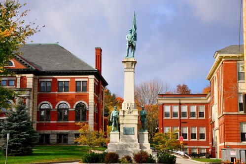 A tall monument with a statue atop, surrounded by trees and historic brick buildings under a cloudy sky.