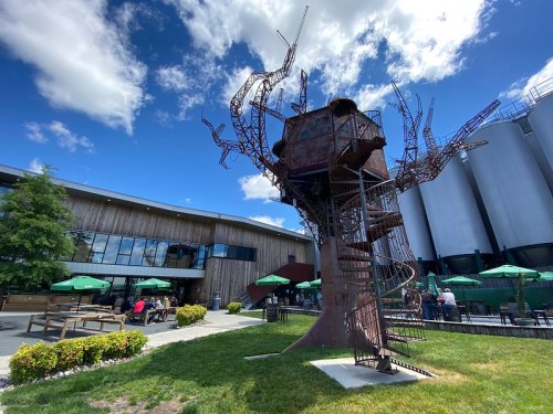 A large metal tree sculpture with a spiral staircase stands in a grassy area near a modern building and outdoor seating.
