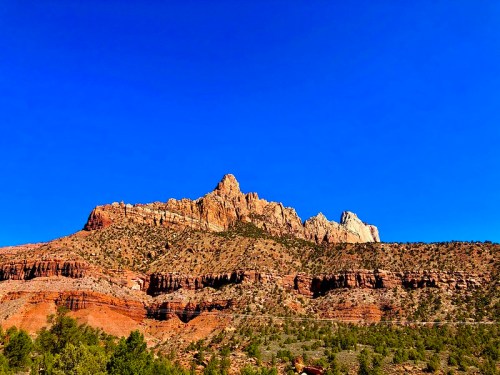 A rugged mountain peak rises against a clear blue sky, surrounded by layered red rock formations and green vegetation.