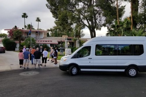 A group of people gathers near a white van in front of a large house surrounded by palm trees.