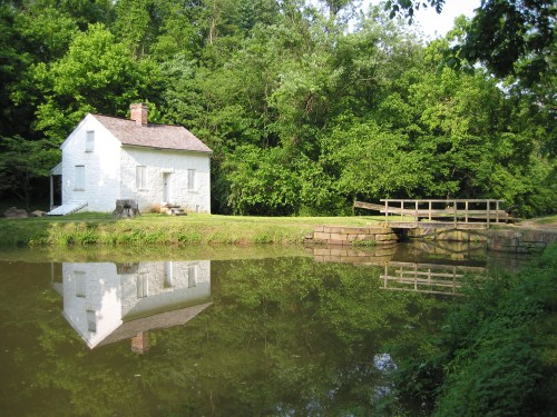A white house beside a calm pond, surrounded by lush green trees, with a small wooden bridge in the foreground.