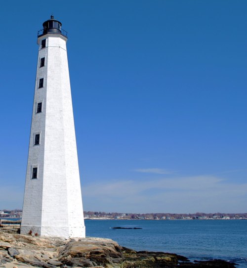A tall white lighthouse stands on rocky shore, overlooking a calm blue sea under a clear sky.