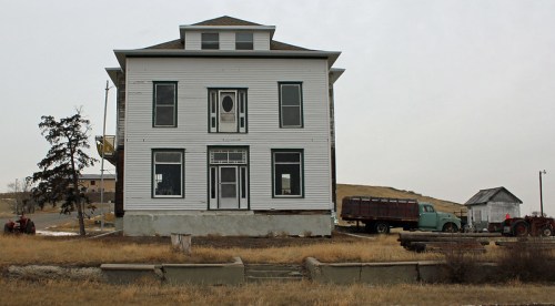 A large, two-story white house with a porch, set against a gray sky and surrounded by dry grass and old vehicles.