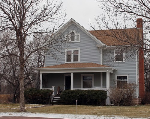 A light blue two-story house with a front porch, surrounded by bare trees and a snowy lawn.