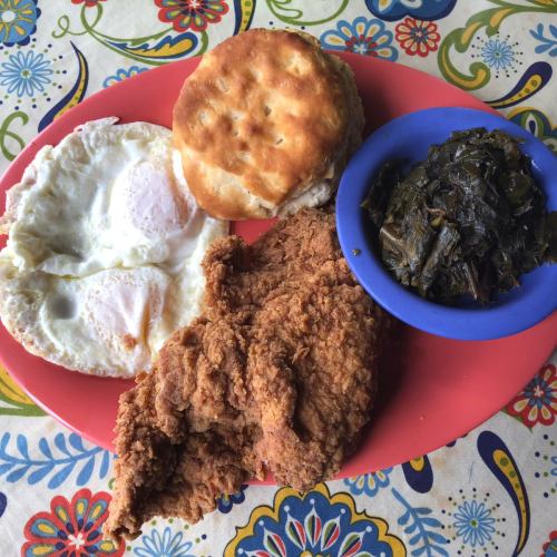 A plate of fried chicken, two sunny-side-up eggs, a biscuit, and a serving of collard greens on a colorful tablecloth.