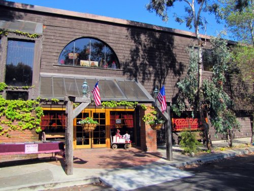 A charming brick building with large windows, adorned with greenery and American flags, set in a sunny outdoor area.