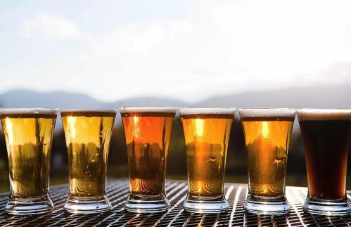 Six glasses of beer in various shades, lined up against a scenic backdrop of mountains and a bright sky.