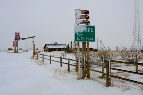 Snow-covered landscape with a sign for Buford, elevation 8000, and nearby gas station and wooden structures.