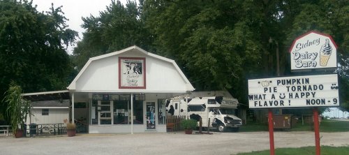A quaint dairy barn with a sign advertising pumpkin pie tornado and happy flavor, surrounded by trees and vehicles.