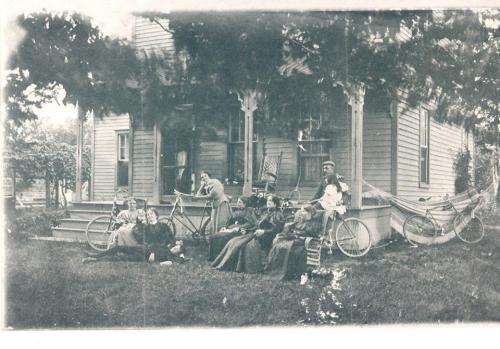 A vintage photo of a group of people with bicycles, sitting on the lawn of a house surrounded by trees.