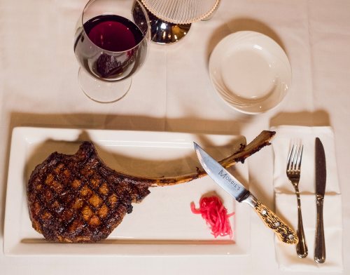 A large grilled tomahawk steak on a white plate, accompanied by a glass of red wine and pickled onions.