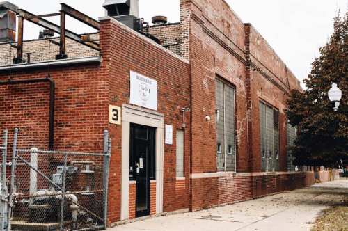 A brick building with large windows and a sign, surrounded by a fence and trees, on a cloudy day.