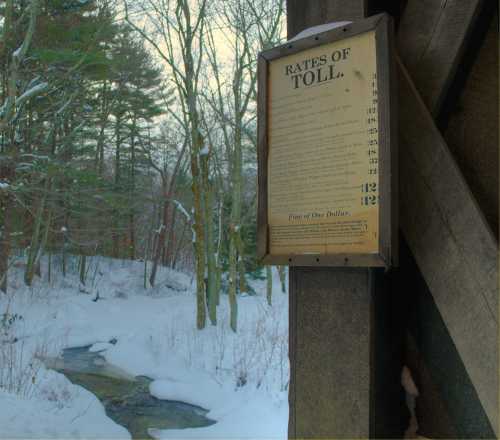 A wooden toll sign hangs beside a snowy landscape with a stream and trees in the background.