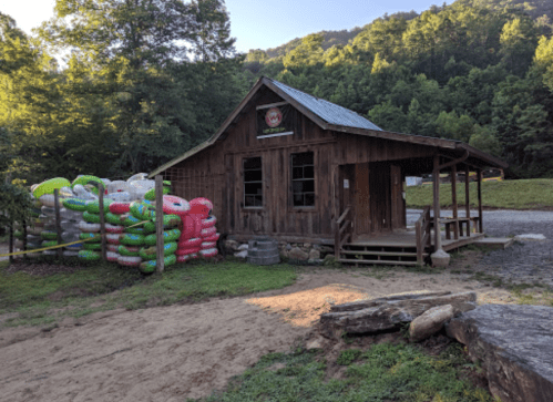 A rustic wooden cabin surrounded by green trees, with colorful inflatable tubes stacked nearby.