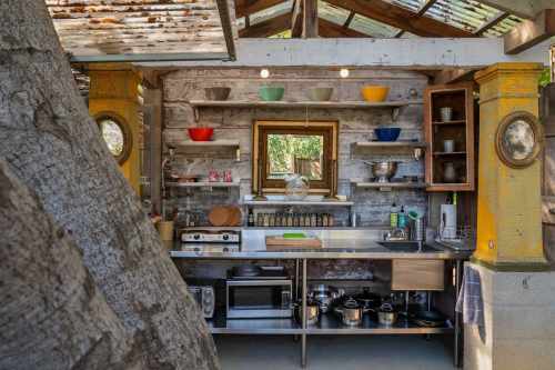 A rustic outdoor kitchen with wooden shelves, colorful bowls, and stainless steel appliances, surrounded by nature.
